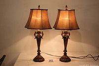 Pair of lamps with tan shades showing the entire lamp, metal base with acanthus leaf designs, and matching finials.