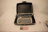 Full view of the Smith Corona Galaxie Deluxe typewriter inside open black hard case on white table, includes a small accessory packet in front.