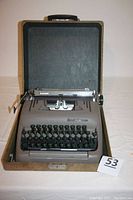Front view of Smith Corona typewriter inside open brown textured carrying case, showing keyboard and paper holding carriage.
