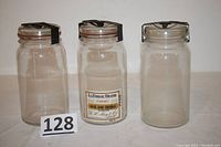Three clear glass mason jars with unusual black metal locking lids, placed side by side on white background with lot number 128.