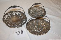 Three ornate metal baskets with detailed openwork grape and leaf motifs and handles shown on a fabric surface with a lot number card.
