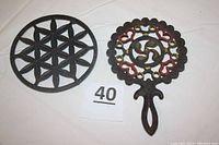 Photo showing two cast iron trivets, one round floral design and one handled with heart and ring pattern with color accents.