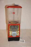 Front view of the vintage gumball machine, showing red top, clear plastic container, and coin mechanism with golden textured background and 25 cent sticker.