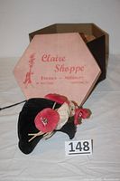 Black velvet vintage hat lying flat next to the original Claire Shoppe hexagonal box, showing floral decoration and shop name on box