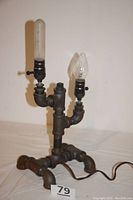 Front view of the vintage steampunk lamp showing design of metal pipes and two light bulbs, one frosted tubular and one clear flame-tip style, with attached power cord.
