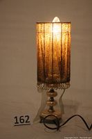 Full view of lamp illuminated showing gold-tone beaded shade with intricate geometric patterns and base with electrical cord.