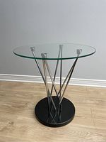 Full view of the end table showing the round glass top, chrome supports and granite base on light wooden floor against grey wall.