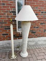 Photo showing two vintage floor lamps placed outdoors on a stone patio against a brick wall: a taller beige floor lamp with a white shade and brass base, and a wider textured plaster lamp with beige and blue coloring and white shade.