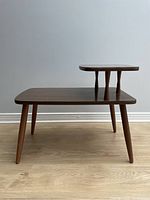Side view of mid century two-tier table showing laminate tiers and four tapered legs on hardwood floor against plain wall.