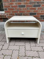 Front view of vintage waterfall style laminate nightstand with one drawer and open shelf, placed on outdoor paved ground against brick wall.