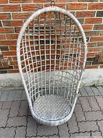 Full view of vintage white wicker hanging chair with woven seat, oval curved frame, placed outdoors against brick wall.