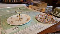 Two single level cake plates on a map background, one with a white base and floral design with a gold-toned handle, the other with a busy floral pattern and silver handle.