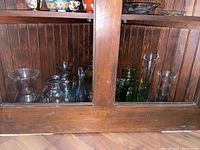 Shelf with multiple clear and colored glass vases and vessels inside wood display case.