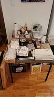 Full view of table with art supplies, palettes, framed prints, boxes and packaged kits densely packed on top.