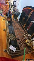 Photo showing fireplace fender, two sets of fireplace tools, brass bucket and bellows next to fireplace