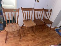 Four antique wooden chairs with spindled backs and worn wooden seats on hardwood floor against a white wall.