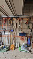 Full view of tools hanging on concrete garage wall including shovels, rakes, hoes, brooms, and extension cords near a bag of soil and coiled hose.