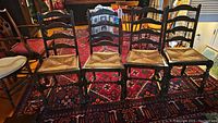 Four wooden ladder back chairs with woven cane seats arranged in a row on a patterned rug, showing the fronts and seats.