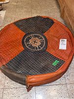 Full view of round leather footstool with two-tone black and brown panels and small white dot patterning, central embroidered cushion, on tiled floor.