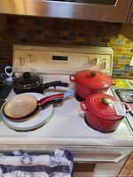 Overall image showing the red cast iron Dutch ovens and assorted frying pans and skillet arranged on a white stove top, providing context for the lot.