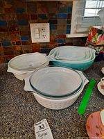 Multiple Pyrex pots and bowls stacked on a kitchen counter, some with floral patterns and clear lids, also nested teal bowls