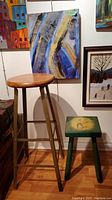 Two stools shown side by side against wall with paintings in background: a tall metal stool with a wooden seat and a shorter green wooden stool with golf artwork on top.