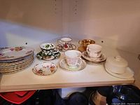 Wide shot of five teacups with matching saucers, additional saucers and Royal Albert Manor Rose patterned plates arranged on shelf.