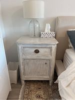 Front view of one night table showing painted finish, drawer and door configuration with lamp and tissue box on top