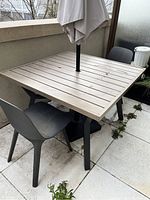 Patio table with wood-look metal slated top and black metal base, shown with two black plastic chairs and closed umbrella in center hole, placed on outdoor balcony floor with plants nearby.
