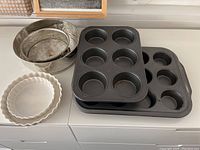 Overview of assorted bakeware including two white ceramic fluted edge pie/quiche pans, two muffin tins (standard and jumbo), and two metal springform pans inside a large stainless steel bowl.