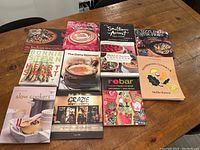 Full view of 11 assorted cookbooks laid out on a wooden table showing front covers and titles.