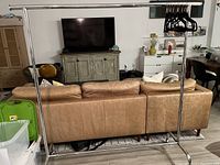 Full front view of the stainless steel rolling clothing rack placed indoors with some clothing hangers on the horizontal bar, showing the overall structure and size relative to a couch and television.
