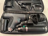 Open black hard carrying case showing PanOptic ophthalmoscope head, otoscope head, lithium ion handle, charger and eyecup attachments