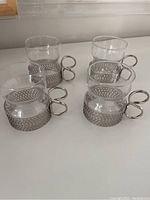 Four clear glass tea glasses each in a silver-tone metal holder with decorative mesh texture and loop handles arranged on white surface.