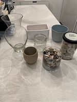 Photo showing various glass and ceramic vases, planters, and containers of river rocks arranged on a white textured fabric.