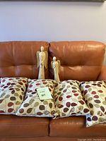Photo showing two wooden angel sculptures and four pillows with a red, brown, and tan leaf pattern on a leather couch.