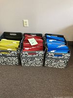 Front view of three soft vinyl file totes with black and white paisley pattern, filled with hanging file folders in yellow, red, and blue.