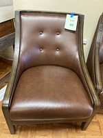 Front view of brown faux leather club chair with wood arms and button tufted backrest.