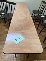 Top-down view of rectangular table surface showing wood-grain laminate finish, surrounded by wooden chairs, with auction lot marker on top.