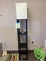 Full view of the tall black shelf tower with white fabric shade on top, placed on carpet near wall outlet with a small decor figure on the middle shelf.