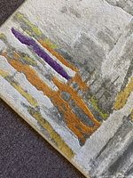 Close-up of rug corner showing orange, purple, yellow, and gray abstract pattern on soft gray and white background.