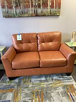 Full view of two-seat orange faux leather loveseat on rug beneath wall art.