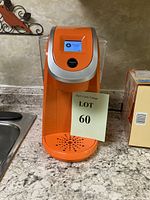 Front angle photo of orange Keurig coffee maker with digital display, on kitchen counter showing the machine and water reservoir.