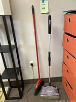 Photo showing Shark electric sweeper and red broom against a wall; charger plugged in.