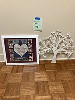 Photo shows the two items side by side on a hardwood floor against a beige wall, a white metal Tree of Life wall hanging on right and a white framed quilt-style print on the left.