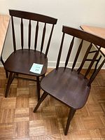 Two brown wooden Windsor-style classroom chairs shown side by side on parquet flooring. Both chairs have spindle backs and contoured seats with a dark brown finish.