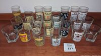 Full set of 25 assorted vintage whiskey and scotch glasses arranged on a wooden surface showing front and side views of branded logos.