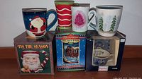 Front view of five Christmas mugs and steins, two mugs feature Santa Claus and candy cane stripes, one a pink tree, and another with Christmas trees. Budweiser holiday stein boxed.