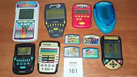 Photo of seven handheld electronic games and four game cartridges on a wooden surface, includes lot number card.
