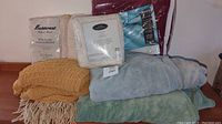 Seven blankets piled including woven, knit, fleece, and packaged blankets in beige, yellow, blue, green and burgundy tones.